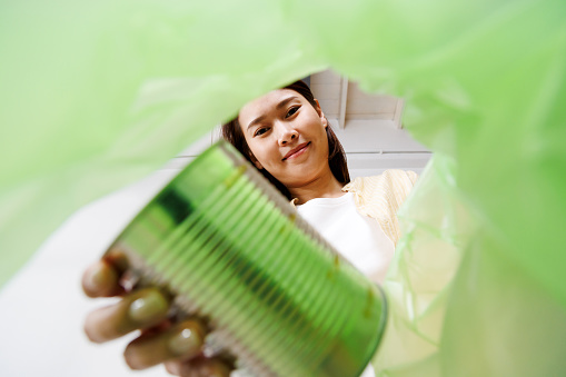 asian woman putting and sorting plastic aluminum can waste to recycle bin POV. Low angle view from inside green recycling bag.