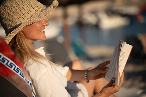 Close-up of a happy blonde woman whit sun hat reading a book at the beach