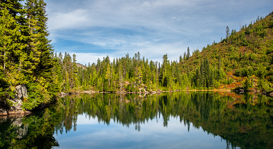 This picturesque image of an alpine lake features a serene reflection of nearby mountains, trees, and the sky on the calm surface of the water, giving a sense of tranquility.  The beauty of this setting lies in the simplicity and natural harmony of the elements and the way they create a captivating scene.  This scene was photographed at Bench Lake in Mount Rainier National Park, Washington State, USA.