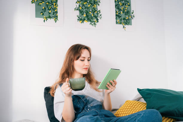 Young woman reading e-book on tablet reader in bed. Female relaxing with hot drink while reading an electronic book in cozy room. Mental health recovery, Personal growth. Hobby, Leisure time alone.