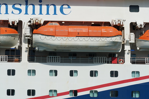 Nassau, Bahamas 12 03 2023: Huge orange life boats secured in the sides of the cruiser passenger vessel with white and blue hull. There is also windows of crew and passenger cabins.