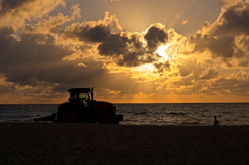 Sunrise on Miami's South Beach brings out the maintenance crew to clean up the beach at first light. Tractor raking the beach in silhouette against warm morning light.
