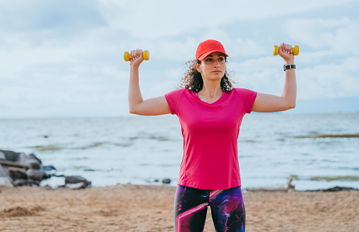 curly-haired young woman in sports clothes does sports on the beach using dumbbells warms up before running. Training fat burning endurance development sports