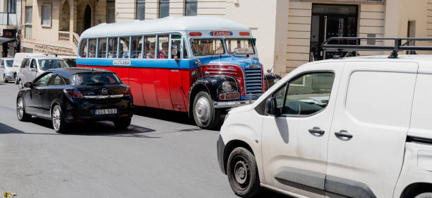 A classic yellow and orange bus is parked in front of ancient stone walls in Malta. The vintage vehicle stands out against the weathered, textured backdrop, evoking a sense of nostalgia and timelessness. Lush green trees provide a pop of color and life to the scene.