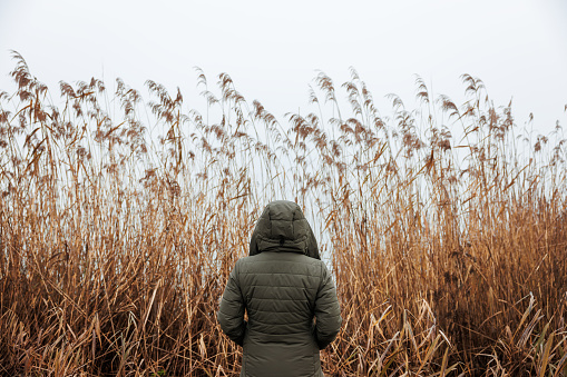 Lonely woman is wearing a hooded coat and standing against reed grass by misty lake. Loneliness concept