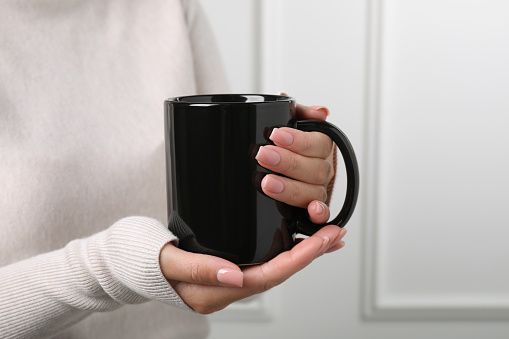 Woman holding black mug indoors, closeup. Mockup for design