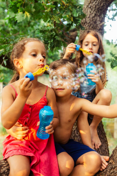 istock Children sitting on a tree and having fun blowing soap bubbles 1902954960