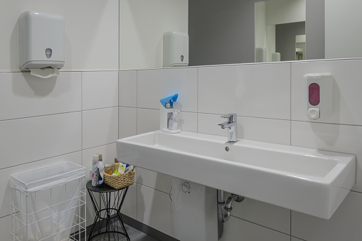 pristine modern bathroom featuring long white sink, hand dryer, and soap dispenser mounted on tiled wall. A basket with cleaning supplies sits beside sink