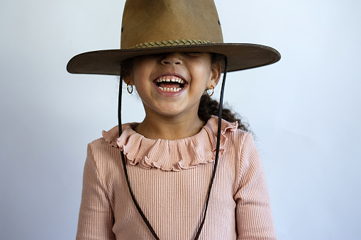 A  young black little girl laughing , wearing earings , a brown hat over her eyes and a pink dress