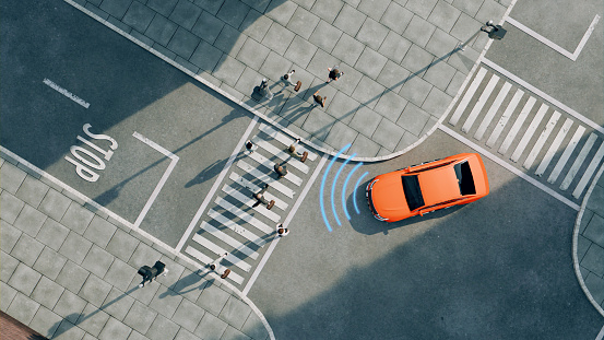 Aerial view of a bright orange car driving on a street. The car is next to a pedestrian crossing and in front of a stop line with the word 