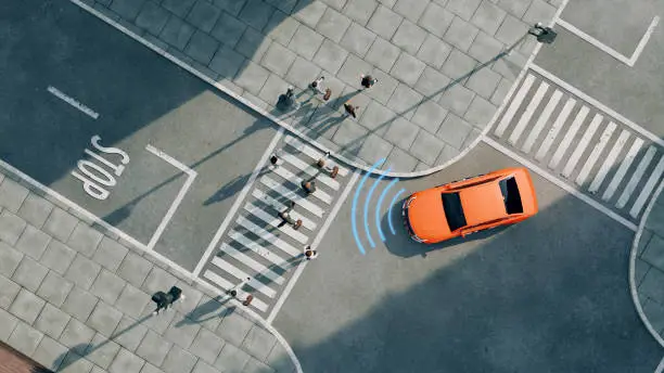 Aerial view of a bright orange car driving on a street. The car is next to a pedestrian crossing and in front of a stop line with the word "Stop" painted on the road. The car uses sensors and radar to detect the pedestrians. Concept of new car technology like autonomous driving.