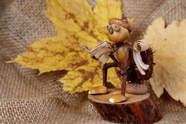 Autumn little man sitting on a stump on a background of yellow leaves and burlap and reading a book. Handmade figurine. Students' works and ideas. There is space for text in the photo.