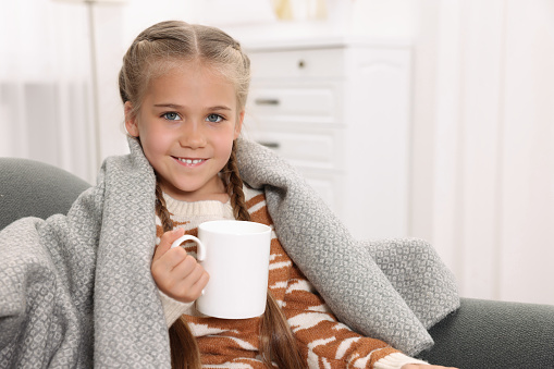 Happy girl under plaid with white ceramic mug on sofa at home, space for text
