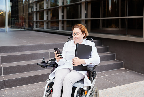 Young businesswoman with a disability sitting in an electric wheelchair. She is using her smartphone. Her disability is the result of spina bifida.