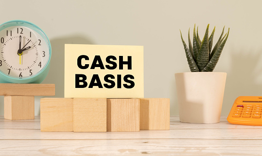 The word cash basis written on a notepaper on business office desktop. Accounting method that recognizes revenues and expenses at the time cash is received or paid out.