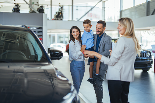 Young happy family enjoying while buying a new car in a showroom. Saleswoman at car dealership center helping family to choose new family vehicle. Family in a car dealership