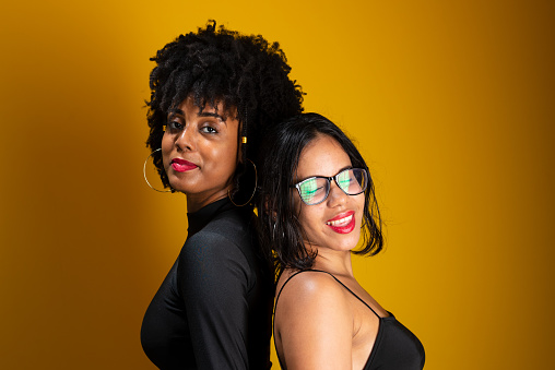 Two friends wearing black clothes, cheerful and happy, playing and posing for a photo against a yellow studio background.