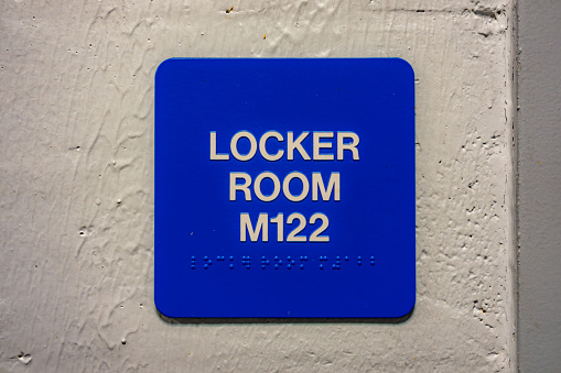 Blue LOCKER ROOM M122 sign with braille, on a white block wall.