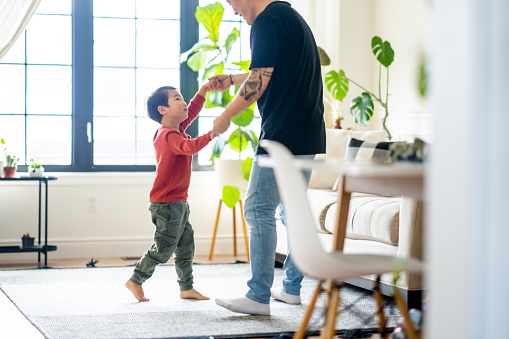 A sweet young boy of Asian decent is seen dancing in the living room with his Dad on a sunny afternoon.  They are both dressed casually as they have fun dancing, flipping and spinning together.