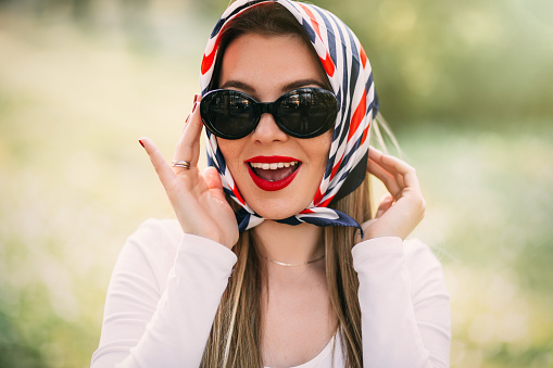 Surprised young girl with red lips in sunglasses and silk scarf wide open mouth looking at camera on green blurred background. Emotions.