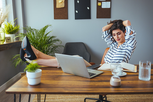 A beautiful business woman in her home sitting in an armchair using a digital laptop, relaxing and using her free time. She put her feet on the table.