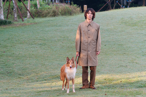 Portrait of a handsome Chinese young man in khaki overcoat with his rough collie dog on green grass field in sunny day, male fashion, cool Asian young man lifestyle, harmony man and pet.
