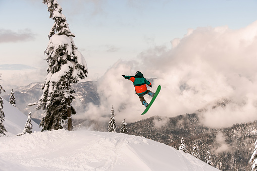 Snowboarder freerider jumping from snow-capped mountain slope on background of trees and cloudy sky. Ski touring in mountains, extreme sport