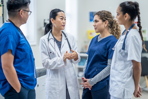 A lead physician has a casual meeting in the hallway as she catches her team up on a patient case and the plan of care.  They are each dressed professionally and are listening attentively.