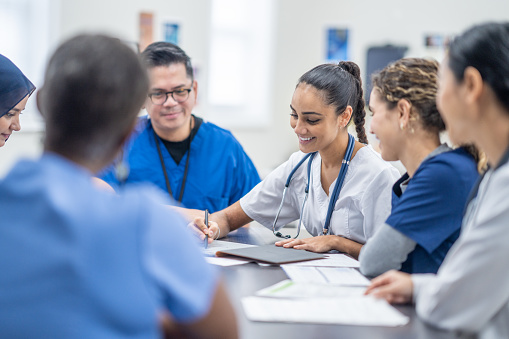 A small group of medical professionals gather around a table as they meet together to discuss patient cases.  They are each dressed professionally and have files scattered between them for reference.