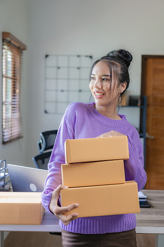 A purple-clad independent Asian woman SME uses a laptop and boxes to receive and review online orders to prepare packages to sell to customers. vertical picture