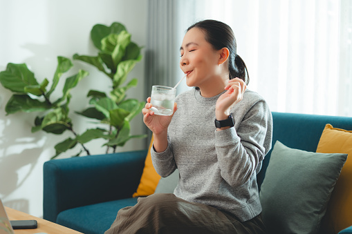 Asian woman taking a break using straw drinking cold water sitting on sofa in living room at home.