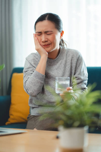 istock Asian woman feels sensitive teeth after drinking cold water and touching her cheek sitting on sofa in living room. 1903716229