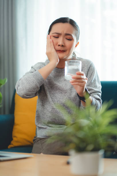 istock Asian woman feels sensitive teeth after drinking cold water and touching her cheek sitting on sofa in living room. 1903717251