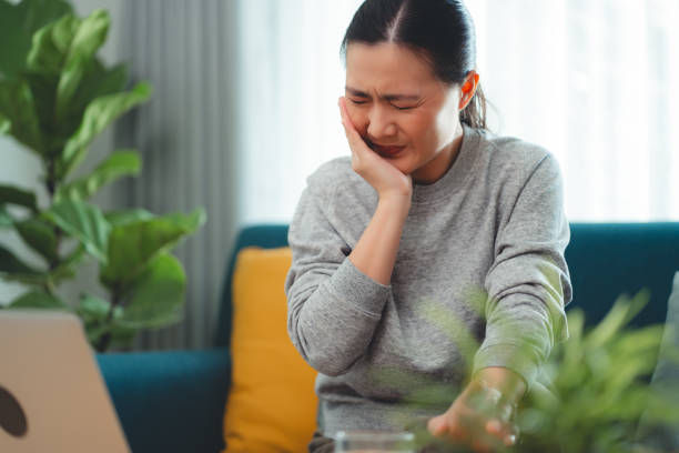 istock Asian woman feels sensitive teeth stop drinking cold water and touching her cheek sitting on sofa in living room. 1903720830