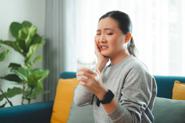 istock Asian woman feels sensitive teeth after drinking cold water and touching her cheek sitting on sofa in living room. 1903724461