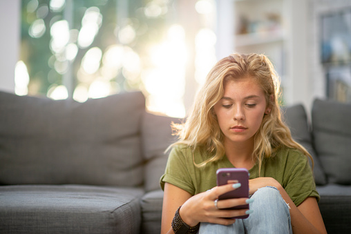 A young teenage girl sits on the floor, leaning up again the sofa as she holds out her cell phone in her hand.  She is dressed casually and is scrolling through her social media while bored at home.