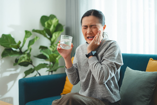 Asian woman feels sensitive teeth after drinking cold water and touching her cheek sitting on sofa in living room at home.