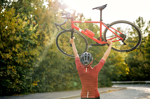 Rear view on Sporty man in sportive clothes, protective helmet and glasses carrying raising bike up after successful competition, posing at camera with red bike outdoors. sport and active lifestyle