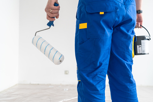 Close-up of a blue-collar worker with a paint can and roller in the empty room prepared for painting