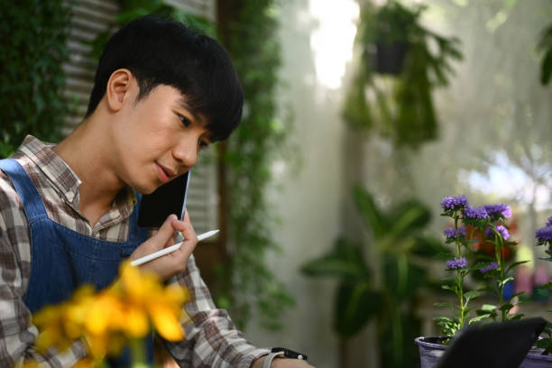 istock Young male florist checking order on digital tablet during conversation with customer on mobile phone. 1903973259