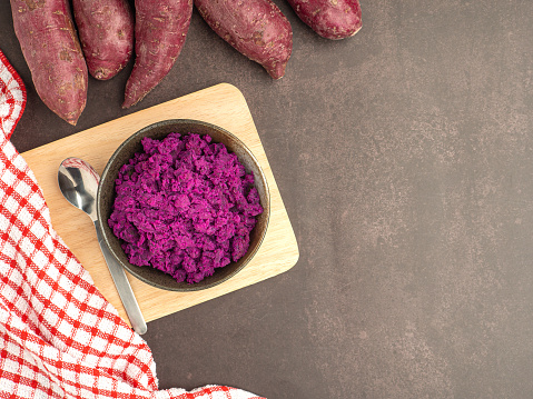 Purple yams pile and mashed purple sweet potatoes on a bowl on a cutting wooden board on a dark brown background. Space for text. Vintage background. Healthy fruits concept