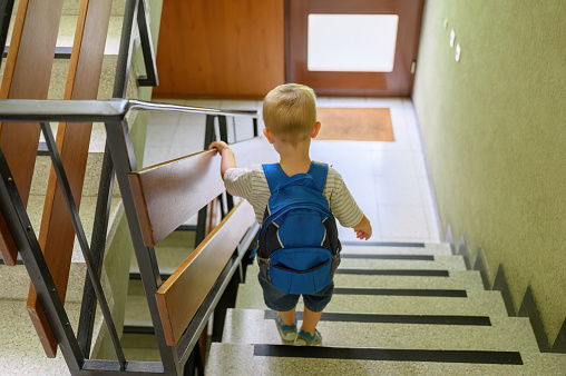 Back view of little boy with blond hair in casual clothes with backpack standing on stairs before going to kindergarten in the morning. family concept