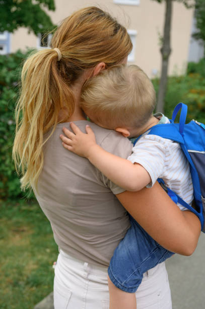 back view of unrecognizable young mother in casual clothes holding son with backpack and hugging on street in the morning back to school concept.