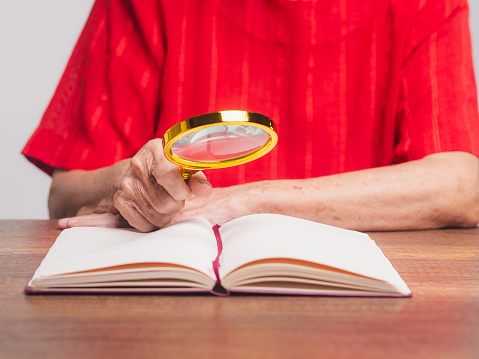 A senior woman is holding a magnifying glass for looking at a book while sitting at the table. Close-up photo. Space for text