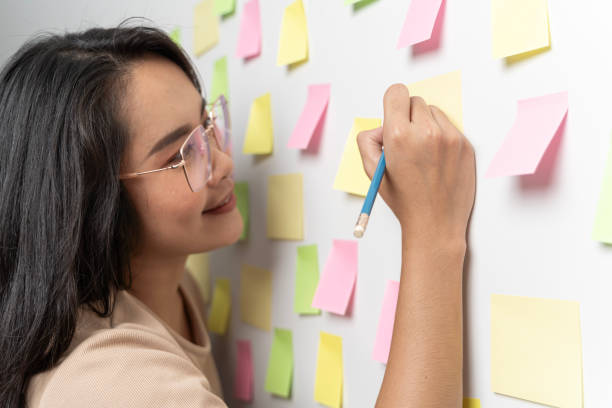 istock Young smiling business woman, leader, consultant, writing creative work on sticky post it notes on glass wall, female coach, project planning. 1904030796