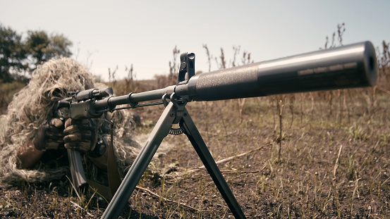 A close-up of a sniper wearing a camouflage suit or grass cloak, waiting and looking through a rifle scope. The sniper aims and shoots. Marksmanship shooting.