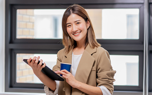 Portrait Asian smart beautiful businesswoman wearing casual business clothes, holding cup of coffee, smiling with happiness, confidence, standing in modern indoor office