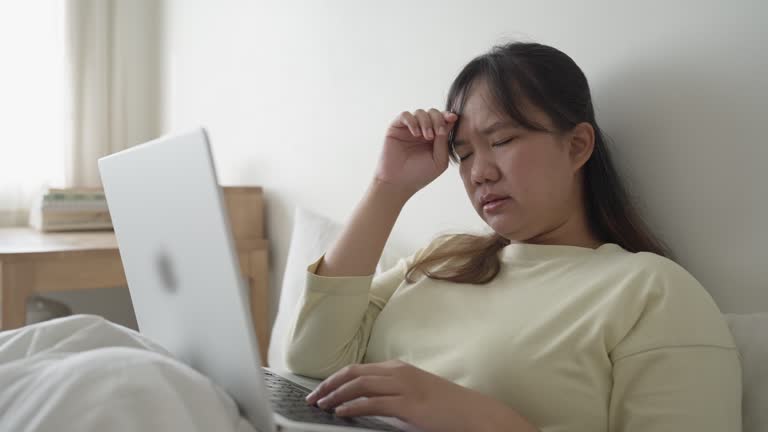 Frustrated Asian woman working using a laptop on the bed to meet deadline