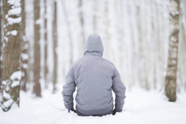 Young adult man sitting on fresh white snow at birch tree forest. Looking far away. Spending time alone in beautiful cold winter day. Back view. Thinking about life. Peaceful atmosphere in nature.