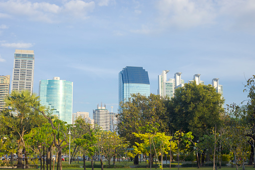 Modern  Bangkok skyline in east of Banjakitti Park seen from northwest of park. In bottom afrea are a parking lot and some people are walking between trees. View from northwestern area of park